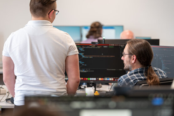 2 Männer im Vordergrund sitzen mit dem Rücken zum Betrachter vor einem Monitor