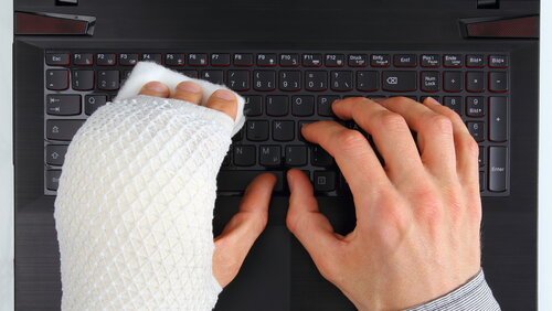 Zwei männliche Hände, die auf einer schwarzen Computertastatur liegen, die linke Hand ist gebrochen und eingegipst.