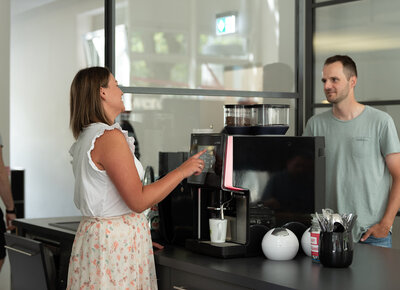 Ein junger Mann und eine junge Frau stehen sich gegenüber an einer Kaffeemaschine und unterhalten sich lächelnd