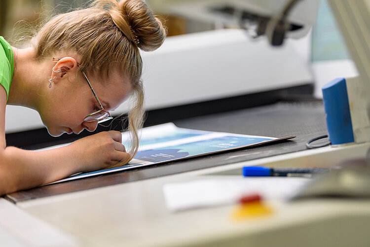 Titel des Bildes Junge Frau schaut mit einem Fadenzähler auf einen Druckbogen
