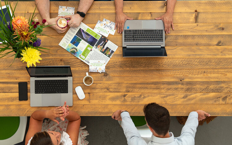 Brainstorming 4 Personen sitzen gemeinsam an einem großen, floral dekoriertem Holztisch und tauschen sich aus, wobei man nur Hände oder den Hinterkopf sieht aus der Vogelperspektive