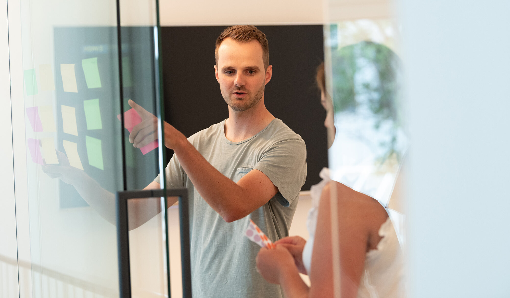 2 Menschen stehen in einem modernen Konferenzraum und hängen Klebezettel an eine Glaswand