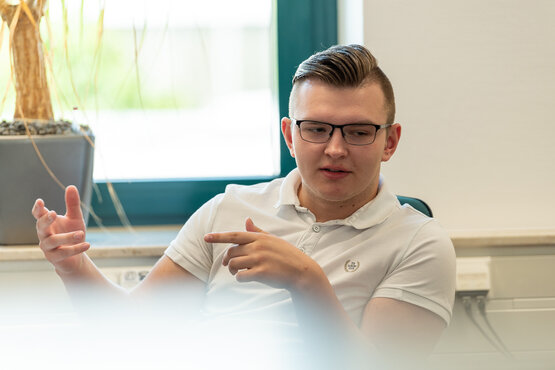 Junger Mann mit Brille sitzt auf Stuhl und erklärt etwas