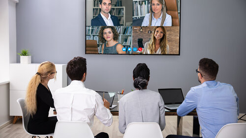 Bild zeigt eine Meetingsituation mit vier Personen in einem Büro und vier Personen die digital per Fernseher zu sehen sind
