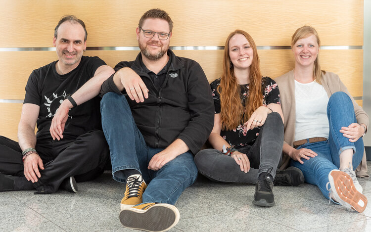 2 Männer und 2 Frauen sitzen nebeneinander auf dem Boden und lächeln in die Kamera