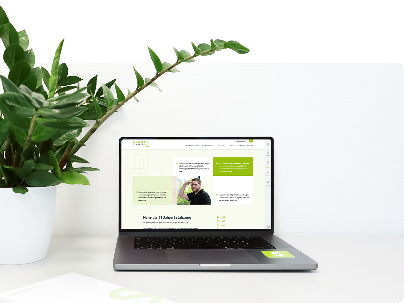 Ein aufgeklappter Laptop steht auf dem Tisch. Links davon steht eine grüne Zimmerpflanze.