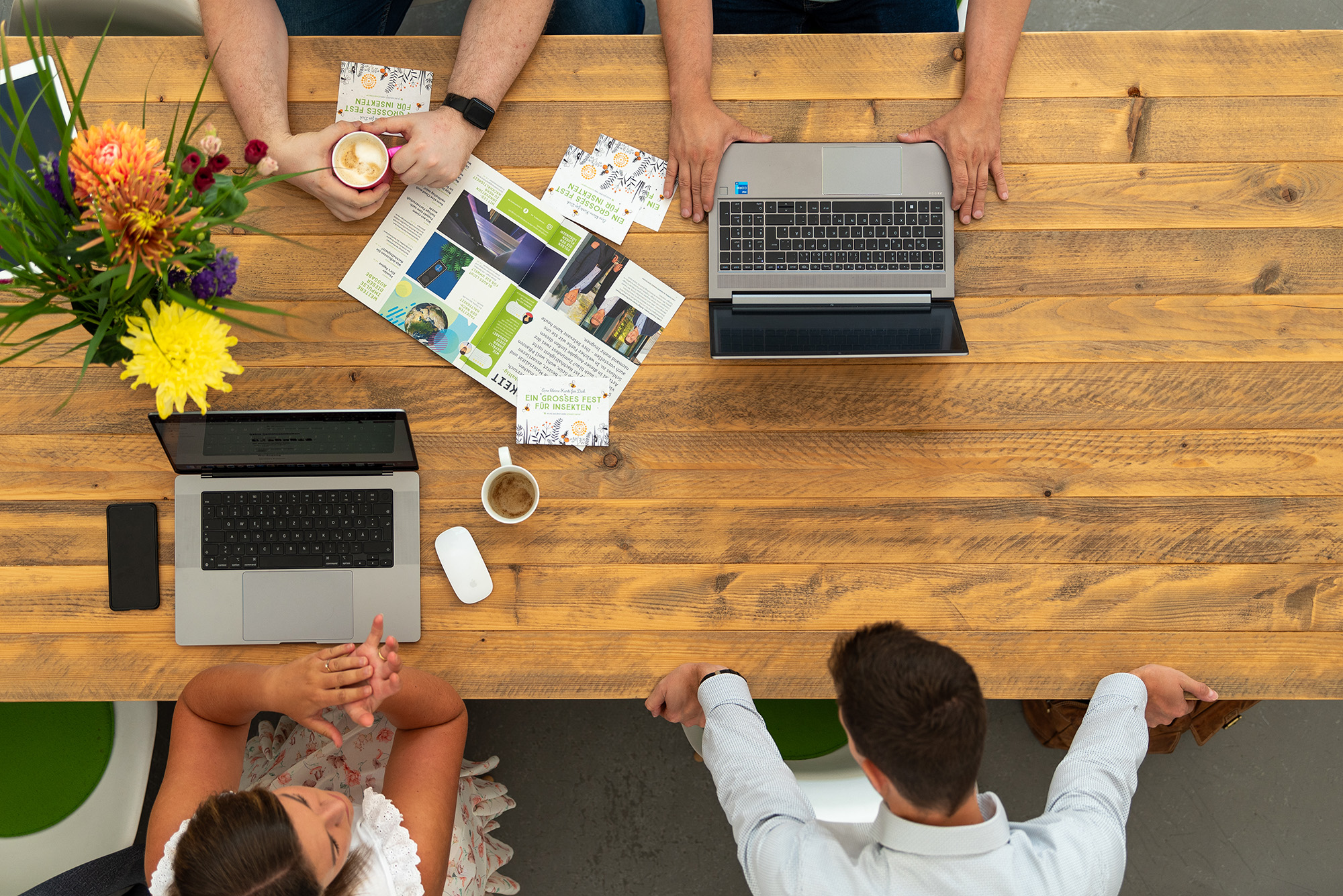 Brainstorming 4 Personen sitzen gemeinsam an einem großen, floral dekoriertem Holztisch und tauschen sich aus, wobei man nur Hände oder den Hinterkopf sieht aus der Vogelperspektive