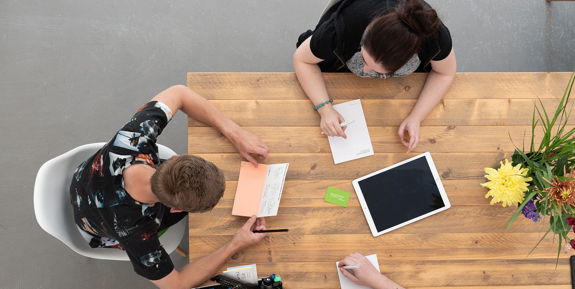 Luftaubhamen von oben: 3 Pesronen an einem Holztisch mit Stiften, Zettel und ipad auf dem Tisch